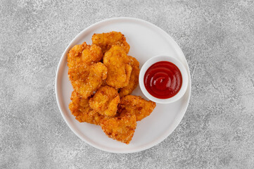 Crispy chicken nuggets with ketchup on a white plate on a gray concrete background.