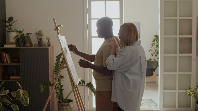 Side view of adult Black man standing at easel in sunlit room and painting while his Caucasian wife embracing him from behind with tenderness and affection, adoring painting