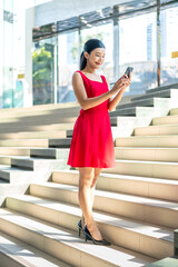 Obraz premium Young Asian Woman in Red Dress Using Phone Indoor a Modern Office Building