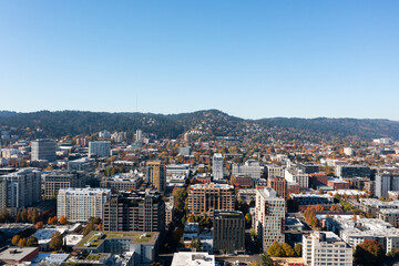 Fototapeta premium Aerial drone view of downtown Portland, Oregon, featuring modern high-rises, urban architecture, and tree-lined streets with Forest Park and the West Hills in the background on a clear autumn day.