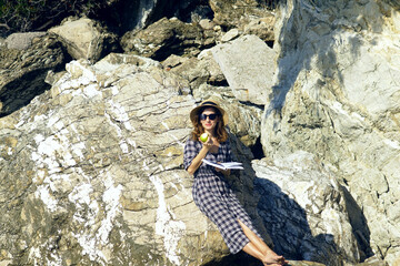 Day off in nature: a young woman in a checkered dress with a hat on her head sits on a sea cliff, reading a book and eating a green apple. A person enjoys relaxing on a wild beach in the Bay of Kotor 
