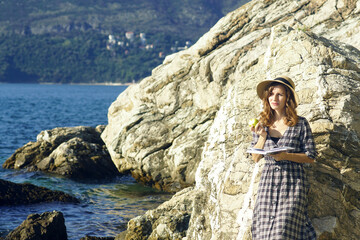 Naklejka premium A person enjoying reading in a picturesque place: a portrait of a young smiling woman standing on the seashore near a rock with a book and a bitten green apple in her hands (Herceg Novi, Montenegro).