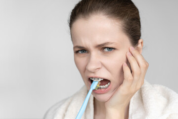 Young woman with braces brushing teeth, holding cheek in pain. Bleeding gums and discomfort shown....