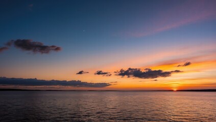 Obraz premium A serene sunset over a calm sea, vibrant orange and yellow hues blending into a soft blue sky, distant dark coastline, gentle waves reflecting the colourful light