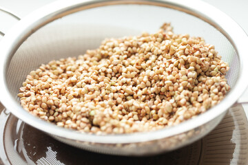 A stainless steel strainer overflows with raw, light brown buckwheat groats, ready for cooking, against a white background.