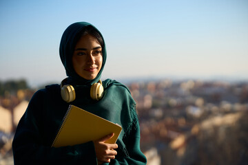 Smiling muslim student holding notebook and looking at city view