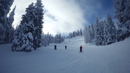 Skiers descend a snowy mountain slope towards a lodge nestled in a snow-covered pine forest under a partly cloudy sky.
