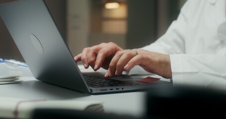 The doctor is typing on a laptop sitting at a table in a medical office, working in a clinic. Close-up of his hands, an unrecognizable person
