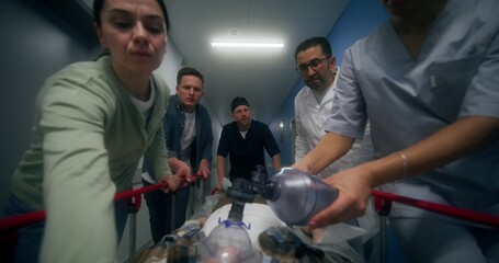 Medical staff and relatives of the victim are running down the hospital corridor carrying the patient on a gurney, giving him oxygen through a mask