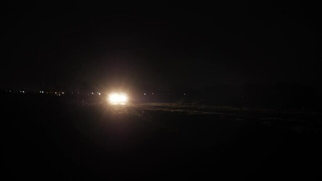 Wide shot of a car approaching in complete darkness, with headlights piercing through the rural night, creating a moody, suspenseful and cinematic atmosphere