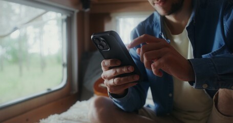 A young man with glasses smiles while using a mobile phone while sitting on a bed in a van. Close-up of his hands