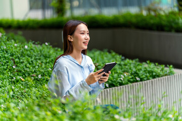 Fototapeta premium Young Asian Businesswoman In Casual Business Outfit Using Tablet Outdoors Sitting In Front a Modern Office Building