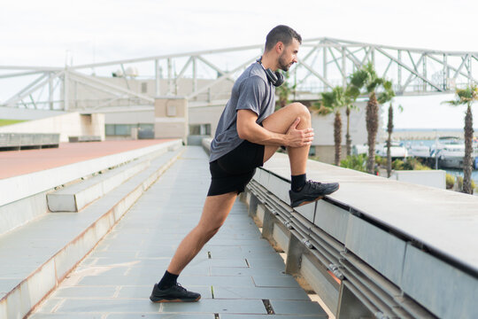 Athletic Man Performing Dynamic Stretching Exercise Outdoors in Stylish Activewear on a Sunny Day to Enhance Flexibility and Prepare for Running or Workout Near the Marina with Palm Trees - Powered by Adobe