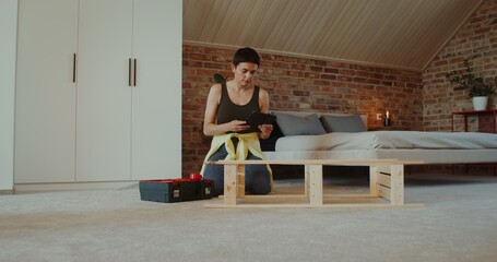 A young woman in casual clothes assembles a rack using a drill, sitting on the floor at home and looking at the tablet screen