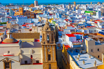 Fototapeta premium Aerial view of city of Cadiz, Spain, dense array of white buildings with flat roofs and prominent church tower in foreground. Vibrant colors of the rooftops and the visible coastline highlight Cadiz