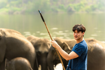 Smiling handsome Asian mahout holding spear weapon taking care elephant in animal conservation