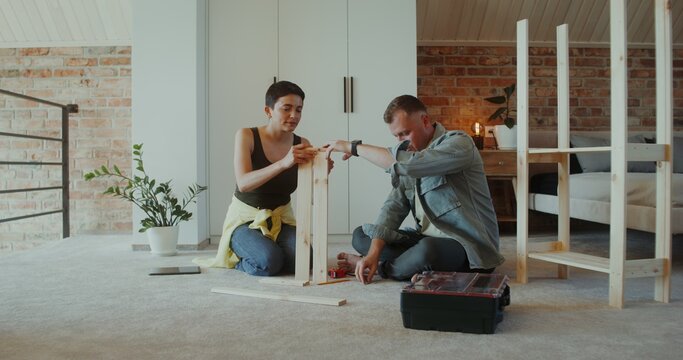 A man and a woman assemble a shelving at home, they use a screwdriver, talk and smile sitting on the floor