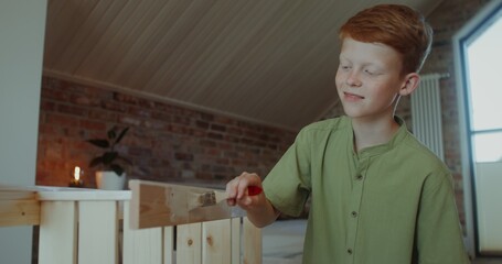 A red-haired boy paints a wooden shelf with a brush while sitting in a stylish home interior