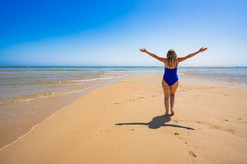 Paradise beach. Beautiful mature woman walking with arms raised up on sandy idyllic beach. Back...