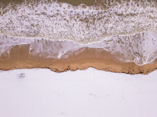An overhead perspective of a winter shoreline where foamy ocean waves meet snowy land. The combination of textures and colors highlights the beauty of seasonal transitions in nature.