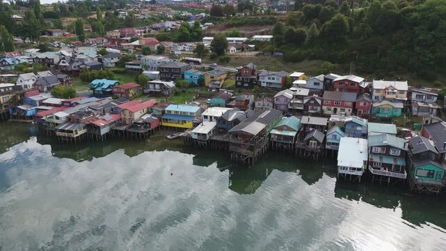 Vibrant stilt houses reflected on waterfront in Castro, Chilo&eacute; Island, surrounded by nature. Aerial Parallax Shot