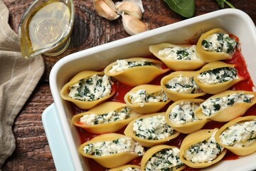 Delicious conchiglie pasta with ricotta cheese, spinach and tomato sauce in baking dish on wooden table, flat lay