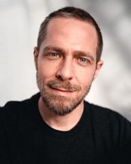 Fototapeta premium Freundliches Close-up-Porträt eines Mannes mit kurzem Bart und blauen Augen. Er trägt ein schwarzes T-Shirt und blickt entspannt in die Kamera. Der unscharfe Hintergrund mit sanften Licht- und Schatte