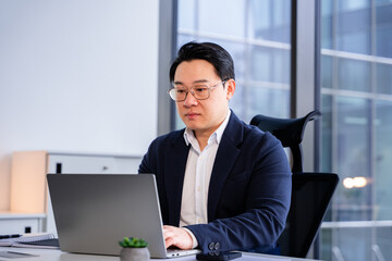 Modern Korean businessman in formal attire working diligently on a laptop in a bright office with large windows, highlighting professional focus and productivity in a contemporary workspace.