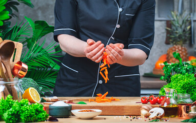 Chef pours chopped carrot from hands. Cozy kitchen with wooden table, kitchenware, vegetables, herbs and ingredients for cooking. Healthy vegan eating, culinary, recipes, food blogging comcept