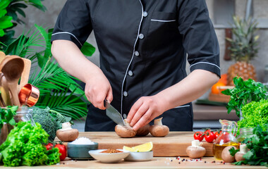 Chef in uniform cuts brown champignon mushrooms. Cozy kitchen with wooden table, kitchenware, vegetables, herbs and ingredients for cooking. Healthy vegan food, culinary, recipes, food blogging