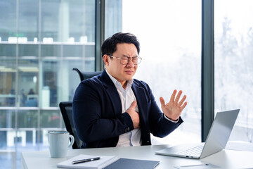 Professional Asian businessman in formal attire holding chest in an office, expressing discomfort...