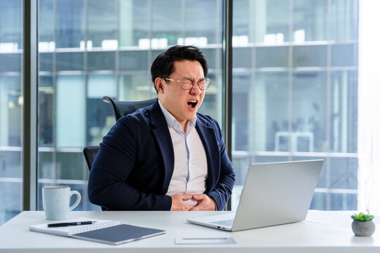 Sick Asian businessman sitting in a modern office space, feeling discomfort while looking at a laptop. Stylishly dressed in business attire, the workspace includes bright lighting and office items.