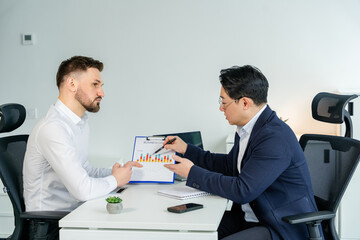 Multinational businessmen discussing financial charts in a modern white office space. Engaged in a workplace presentation, formal business attire. The environment is calm, focused, collaborative.