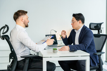 Formal business discussion at modern office between two professionals, reviewing reports. Collaborative work environment displaying productivity, focus, and professional attire at a corporate setting.