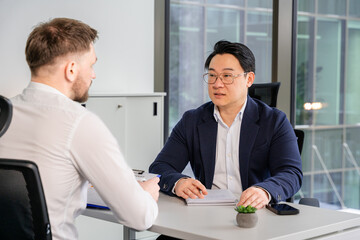 Professional business discussion in a modern office setting with two individuals in formal attire near large windows and neutral colors, underlining the work environment and collaboration focus.