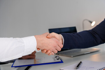 Two professionals shaking hands in an office setting, symbolizing agreement, teamwork, and collaboration, with documents and a laptop in background, illustrating business interactions and cooperation.