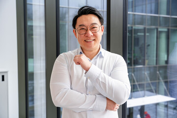 Fototapeta premium Successful Asian male professional wearing glasses standing confidently in a modern office setting, expressing positivity and professionalism in a white shirt with large windows in the background.