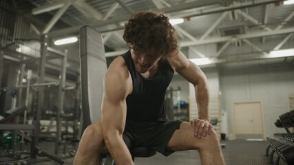 Strong man wearing black tank top performing one-arm bicep curls with dumbbell while sitting on bench in modern gym, surrounded by weightlifting equipment and machines - Powered by Adobe
