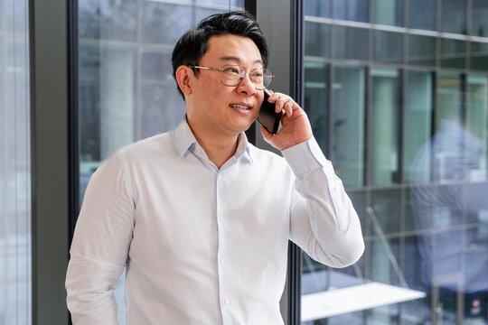 Confident businessman wearing glasses and white shirt talking on a smartphone by a window in a modern office. Depicts communication, professionalism, and a productive work environment.
