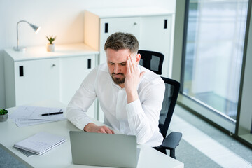 Fototapeta premium Professional businessman in a white shirt, seated indoors at an office desk, appearing under stress while working on a laptop. Modern workplace environment with documents and bright glass windows.