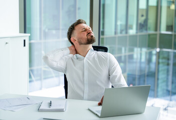 Professional businessman in a white shirt, sitting at an office desk with a laptop and papers,...