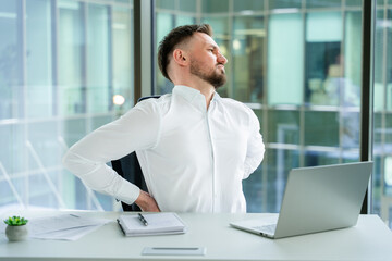 Professional businessman in white shirt sitting in an office, stretching back to relieve discomfort...