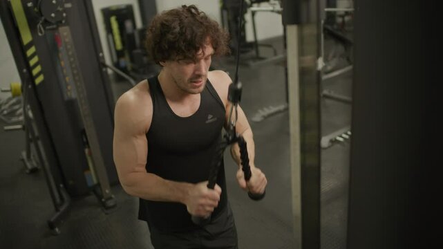Muscular man in sportswear performing triceps pushdown exercise using cable machine during workout in modern gym. High angle view