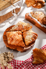 Golden flaky croissants with almond toppings displayed on a rustic wooden board, set on a white tablecloth with a red checkered napkin, creating a cozy and inviting bakery atmosphere