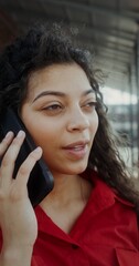 A young woman in casual clothes smiles while talking on a mobile phone and drinking mojito standing on the balcony. The wind blows her hair