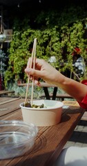 A young woman in casual clothes eats Asian food out of a box using chopsticks while sitting at a table on the cafe terrace, hands close-up, no face