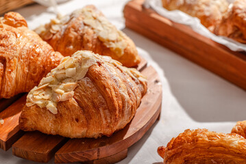 Golden flaky croissants with almond toppings displayed on a rustic wooden board, set on a white tablecloth with a red checkered napkin, creating a cozy and inviting bakery atmosphere