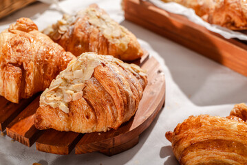 Golden flaky croissants with almond toppings displayed on a rustic wooden board, set on a white tablecloth with a red checkered napkin, creating a cozy and inviting bakery atmosphere