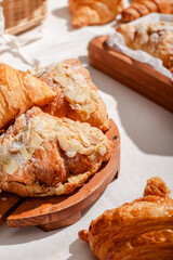 Golden flaky croissants with almond toppings displayed on a rustic wooden board, set on a white tablecloth with a red checkered napkin, creating a cozy and inviting bakery atmosphere