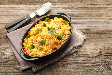 Tasty rice with vegetables in baking dish and cutlery on wooden table, space for text